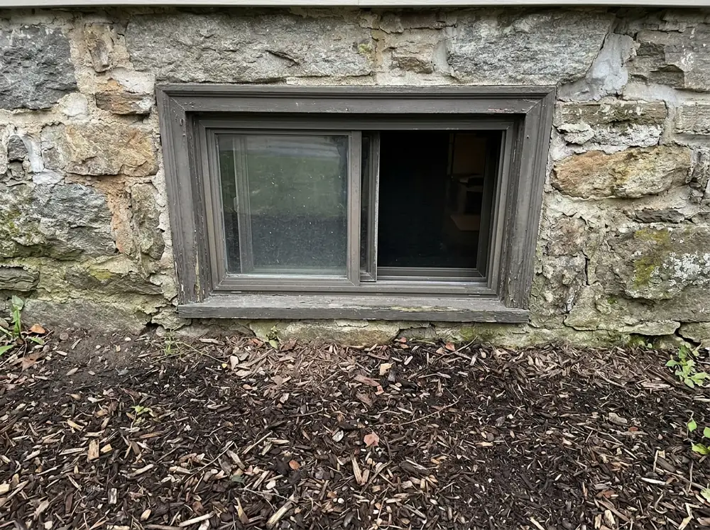 Horizontal sliding basement window with one panel open, set into a stone and mortar foundation wall just above the soil line