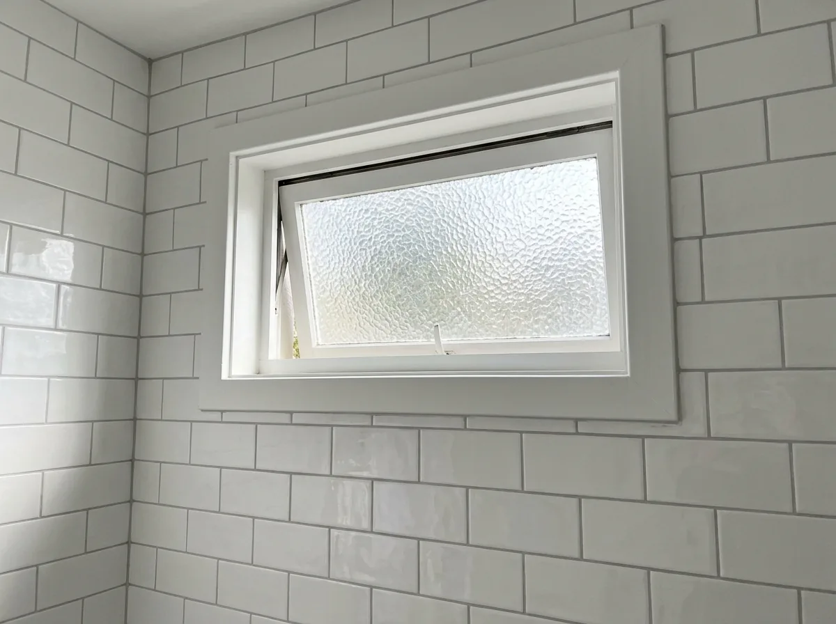 Small awning window set high on a white tiled bathroom wall, fitted with textured obscure glass that blurs the view outside